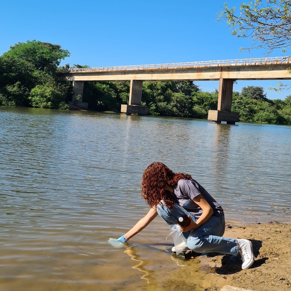 Uma pessoa com cabelo cacheado coleta uma amostra de água na margem de um rio. Ao fundo, é possível ver uma ponte sobre o rio cercada por vegetação verde.